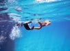 A person in swim trunks swimming underwater, partially submerged, with bubbles rising near their legs in a clear blue pool.