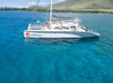 A white catamaran named "Alihilani" floats on clear blue water near a green, mountainous shoreline under a partly cloudy sky.
