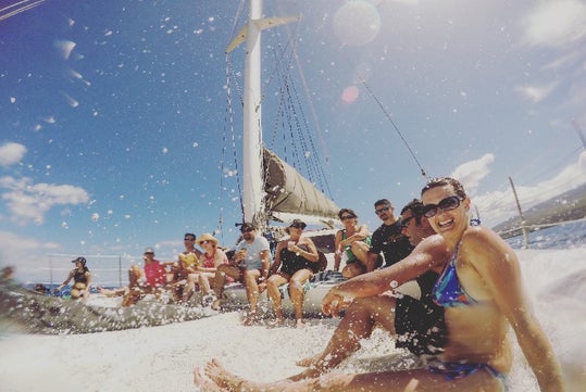 A group of people sit on a sailboat in sunny weather, smiling and getting splashed by water as they enjoy the ride.