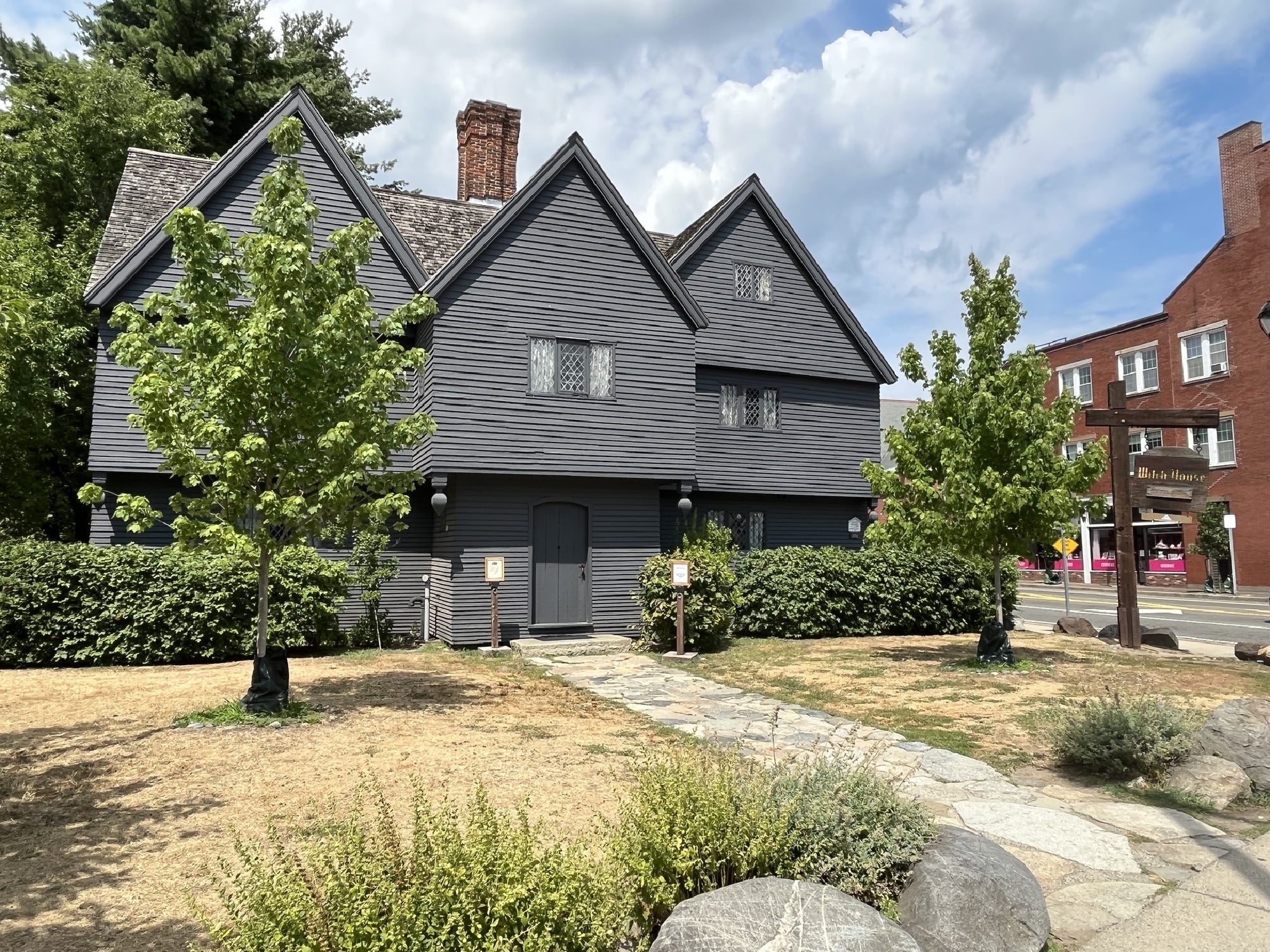 A gray, two-story, historic wooden house with diamond-paned windows stands near a sidewalk, surrounded by shrubs and a partially dry lawn under a partly cloudy sky.