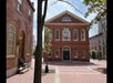 A red-brick town hall building with arched windows stands in a quiet, sunlit plaza lined with brick pathways and a few trees.