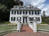 A large white colonial-style house with black shutters, three dormer windows, twin chimneys, and a white picket fence with a brick walkway leading to the front door.
