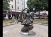 Bronze statue of a woman in a dress sitting on a crescent moon, located in an outdoor plaza with shops and trees in the background.