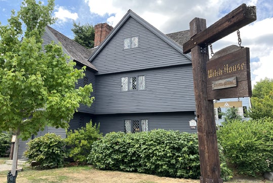 A dark gray wooden house with diamond-paned windows stands behind a wooden sign that reads "Witch House." Shrubs and a tree are in the front yard.