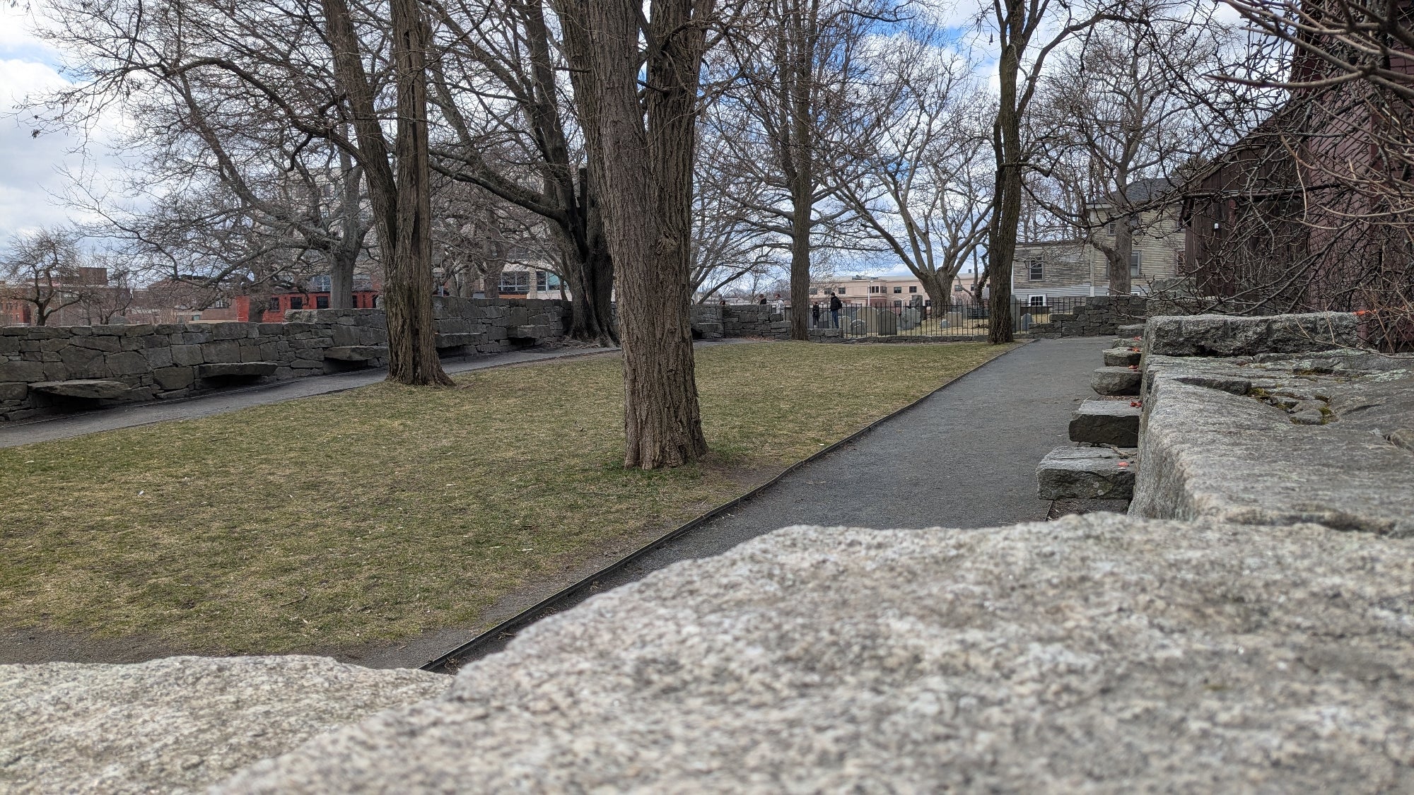 A paved path runs through a small park with leafless trees, stone walls, and buildings visible in the background under a cloudy sky.