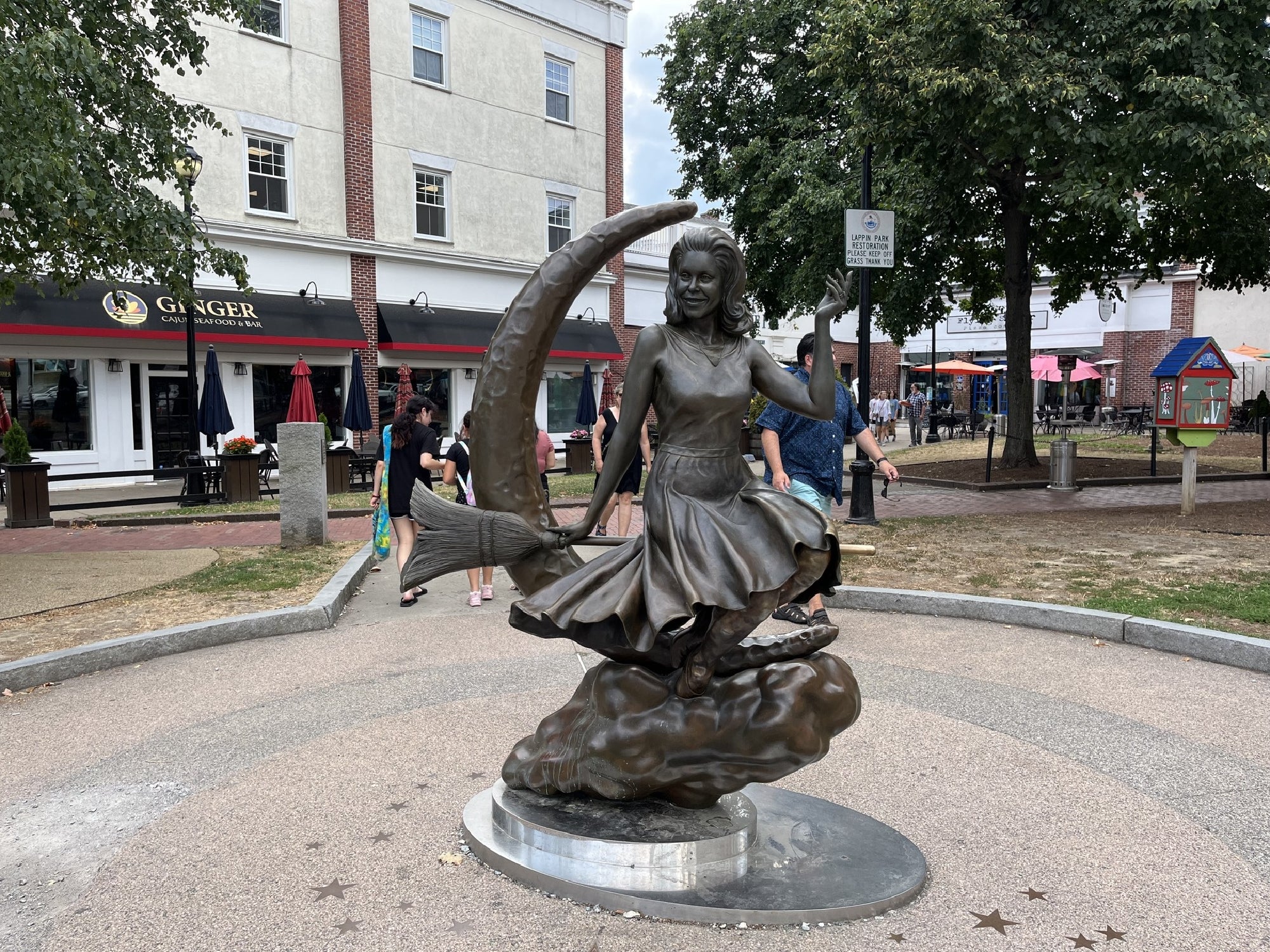 Bronze statue of a girl riding a fish, holding a crescent moon, set in a circular plaza with people, trees, and buildings in the background.
