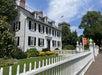 A large white colonial-style house with black shutters and a picket fence, surrounded by greenery and flowers, under a blue sky with clouds. An American flag is visible in the background.