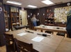 Classroom with wooden tables, chairs, and various art supplies; a person stands near the back wall, which is decorated with notes and crafts.