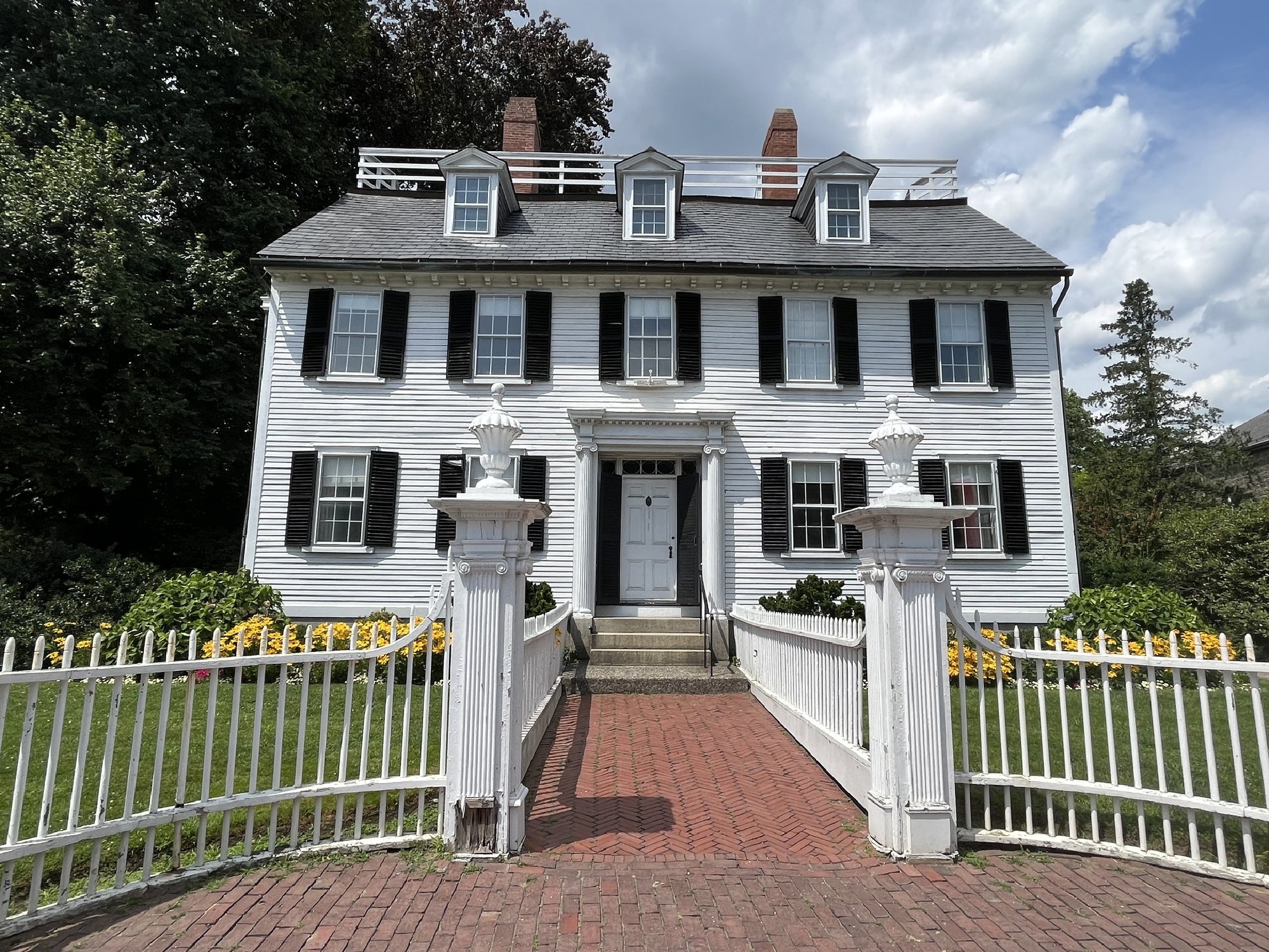 A large white colonial-style house with black shutters, a centered front door, three dormer windows, white picket fence, and brick walkway under a partly cloudy sky.