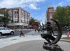 A bronze statue of a woman sitting on a crescent moon faces a crosswalk in a busy downtown area with brick buildings and pedestrians.