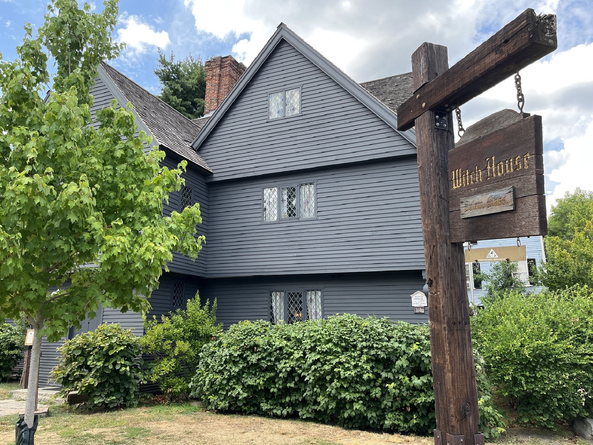 A gray wooden house with diamond-paned windows, leafy bushes in front, and a wooden sign reading "Witch House" stands under a partly cloudy sky.