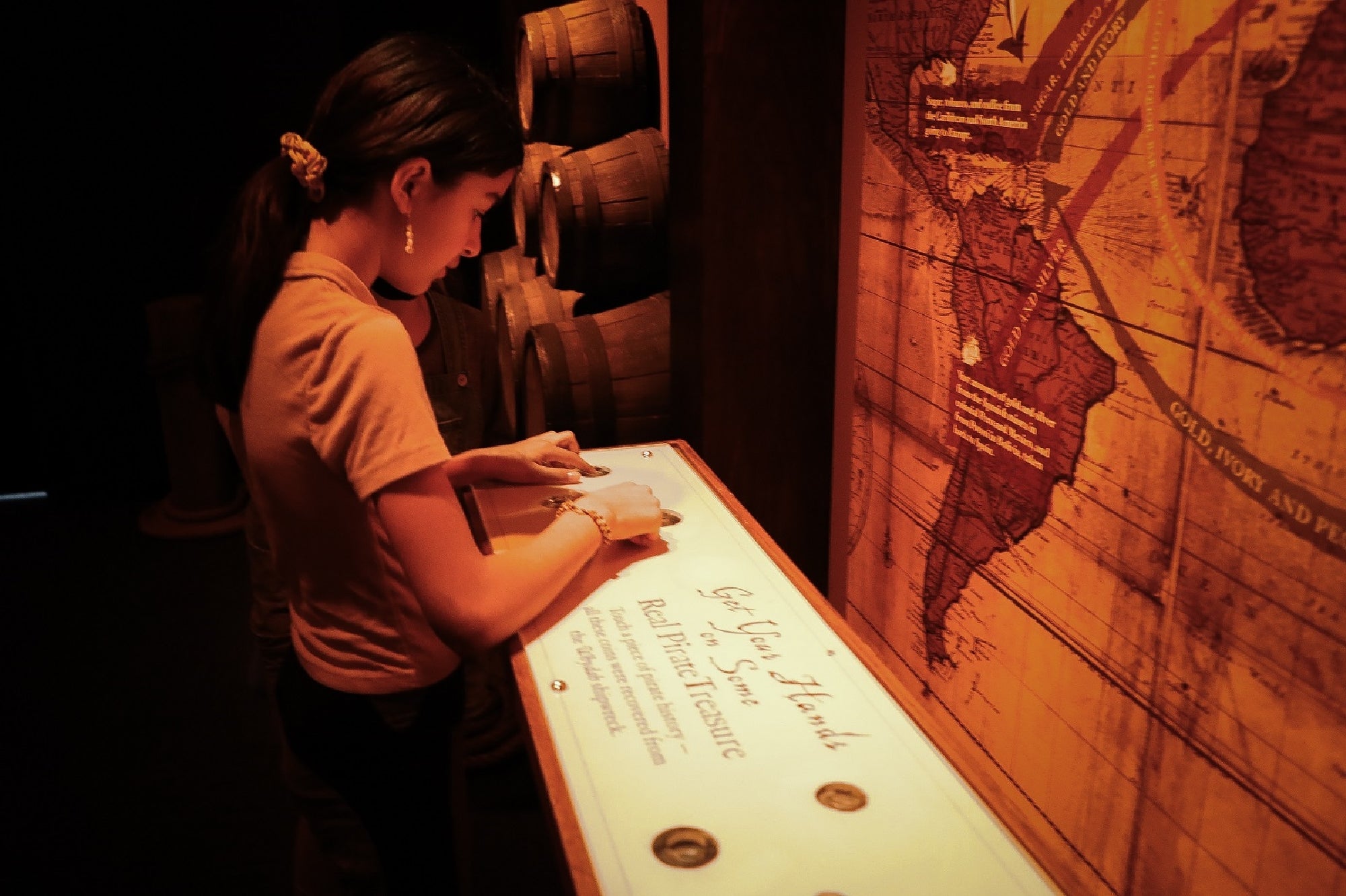 A woman examines replica coins at an interactive exhibit in a dimly lit museum, beside a large vintage map on the wall.