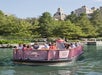 A group of people are touring on a purple riverboat in a lush, scenic area with trees and a large building in the background.