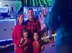 A family stands together and smiles for a photo in front of a brightly lit vehicle decorated with colorful holiday lights at night.