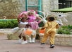 Two children in animal costumes holding orange buckets approach an adult dressed in pink with feathers, standing outdoors near stone walls and plants.