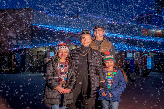 A family of four stands outside at night, surrounded by snow. They are bundled in winter coats and hats with decorative lights in the background.