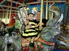 A man and a young child in a bee costume sit on an elephant carousel ride, smiling at the camera. A carousel rooster is visible in the background.