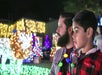 An adult and child wearing holiday light necklaces look at a brightly lit lion decoration and colorful holiday lights at night.