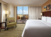 Hotel room with a large bed, armchair, and a side table. Glass doors lead to a balcony with outdoor furniture and a view of the cityscape.
