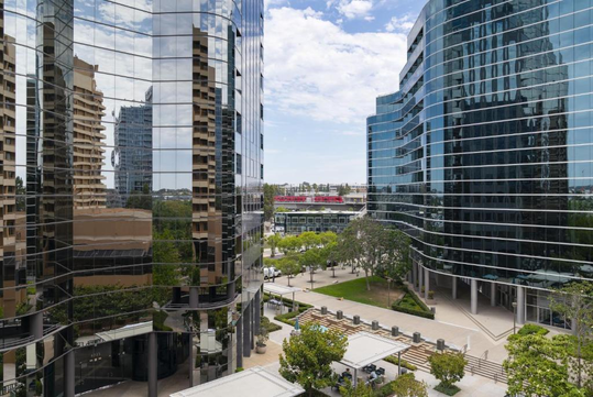 View of modern glass office buildings with reflections, surrounded by trees and a clear sky. A distant stadium is visible between the buildings.