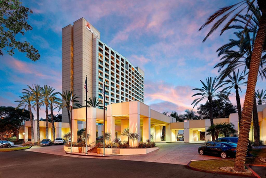 A multi-story hotel with a modern design and palm trees in the foreground, set against a sunset sky.