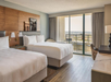 A hotel room with two double beds, modern decor, a desk, a lamp, and a balcony with a view of the city skyline through sliding glass doors.