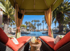 View from inside a poolside cabana with orange cushions, looking out toward a swimming pool, palm trees, and clear blue sky; a table holds drinks and a fruit bowl.