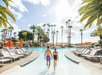 A man and woman in swimwear walk hand in hand into a large outdoor pool surrounded by lounge chairs and palm trees under a partly cloudy sky.