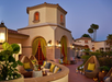 A Mediterranean-style villa with arched doorways, outdoor seating, lanterns, plants, and decorative lighting under a clear evening sky.