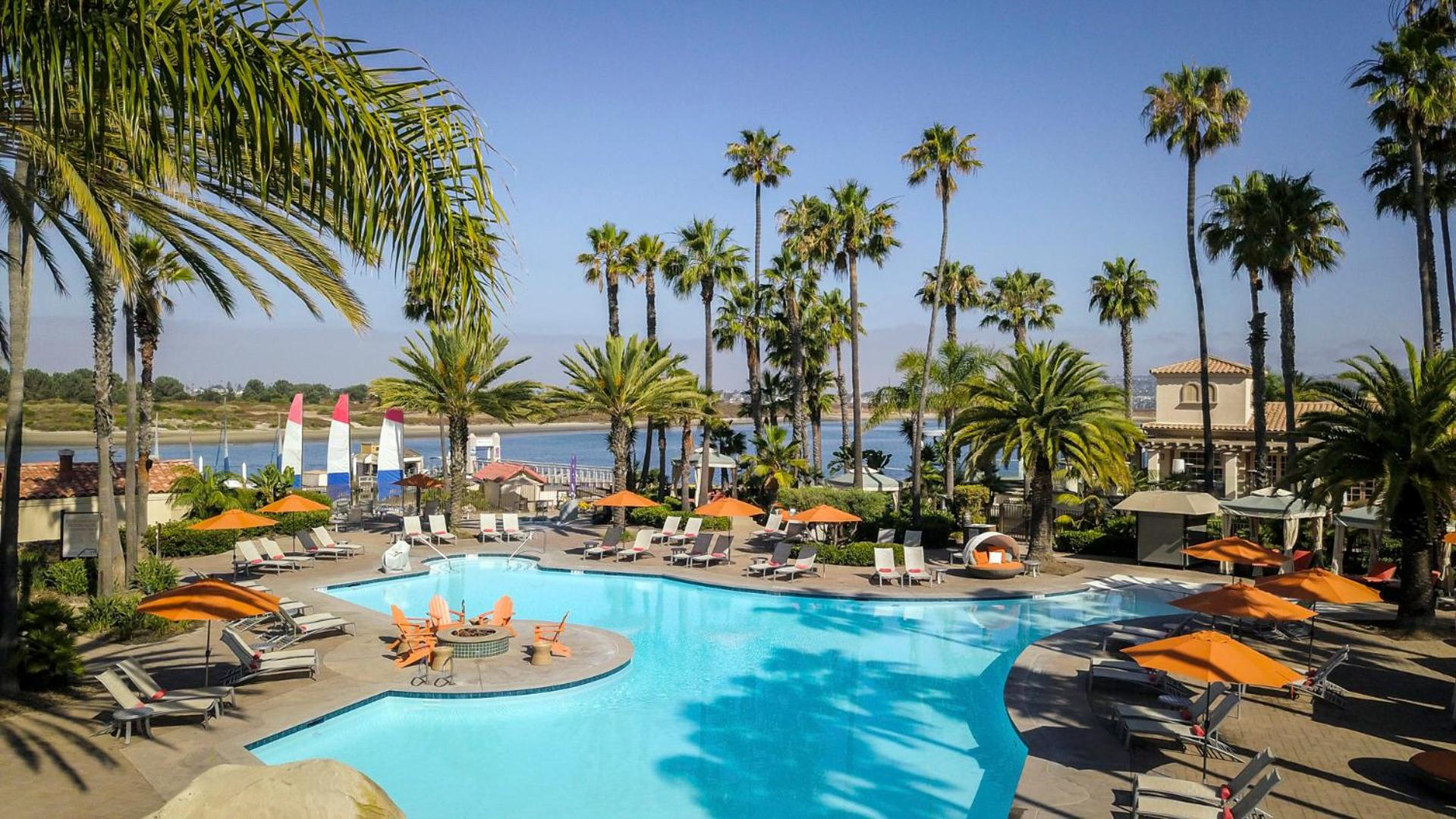 Outdoor pool area with lounge chairs, umbrellas, and palm trees overlooking a body of water under a clear blue sky.