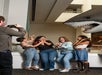 Four women pose with playful, surprised expressions in front of a large shark model while a man takes their photo in a museum setting.