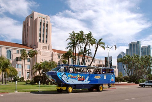 A blue amphibious tour bus with passengers drives past a large historic building surrounded by palm trees and modern skyscrapers under a partly cloudy sky.