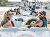 Four people wearing sunglasses and life jackets sit in small white motorboats docked at a marina, smiling and looking toward the camera. Boats and water are in the background.