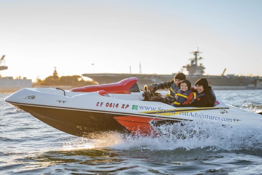 Three people wearing life jackets ride a small speedboat on the water, with a large ship in the background under a clear sky.