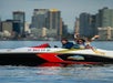 Two people ride a small motorboat on the water, with tall city buildings in the background. One person is waving with both hands.