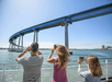 Three tourists standing near the railing of a cruise boat taking photos on a sunny day with a bridge in the background.