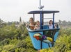 Three people ride in a blue cable car decorated with a painted bird, moving above trees with buildings visible in the background.