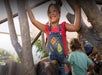 A child wearing a red shirt and denim overalls with patches plays on a tree structure with other children around.