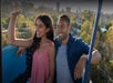 A smiling woman and man enjoy a ride on an open cable car, with trees and distant buildings in the background.