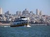 A ferry navigates across the water with a city skyline in the background under a clear blue sky.