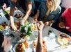 People seated around a table share a meal, reaching for bacon and other breakfast foods on white plates under natural sunlight.