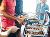 Three people serving themselves food from a buffet table with trays of hot dishes, holding plates and utensils.