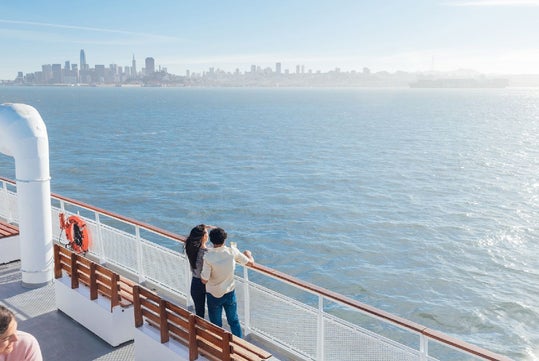 Two people stand by the railing on a ferry with a city skyline visible in the background across the water on a clear day.