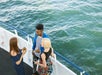 Three people stand on a boat deck holding drinks and talking, with the ocean visible in the background.