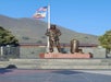 Bronze statue of a military figure standing beside a large artillery shell on a pedestal, with American and California flags waving in the background. Hills and trees are visible.