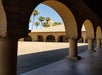 Stone archway leading to an open courtyard with palm trees and a bell tower visible in the background under a clear blue sky.