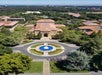Aerial view of a campus with a central circular fountain, surrounded by trees and buildings with red-tiled roofs. A group of people is gathered near the fountain.
