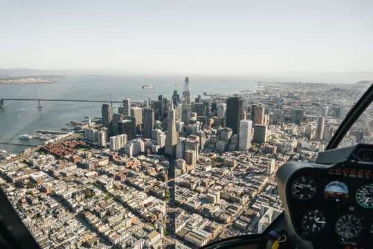 Aerial view of a cityscape with tall skyscrapers by the waterfront. A bridge extends over the water. The image is taken from inside a cockpit, with control dials visible in the foreground.