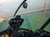 View from inside a helicopter cockpit flying over a body of water, with a red suspension bridge prominently visible below.