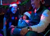 Three people wearing Santa hats sit together in a decorated vehicle, smiling and illuminated by colorful holiday lights.